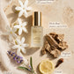 A bottle of Cleo VII Jasmine Perfume Oil is styled in a flat lay with labeled botanical ingredients including white flowers, lavender, turmeric, lemon, and a nut, on a beige stone background.